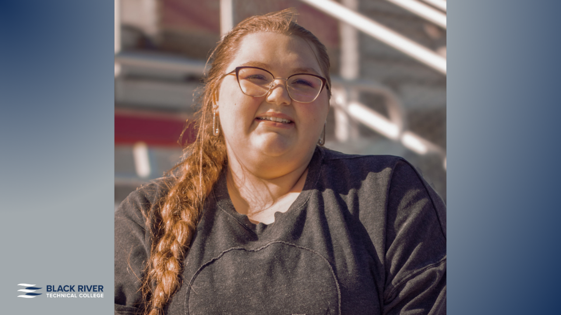 A young woman with long red hair in a braid smiles outdoors, wearing glasses and a dark sweatshirt, with bleachers visible in the background.